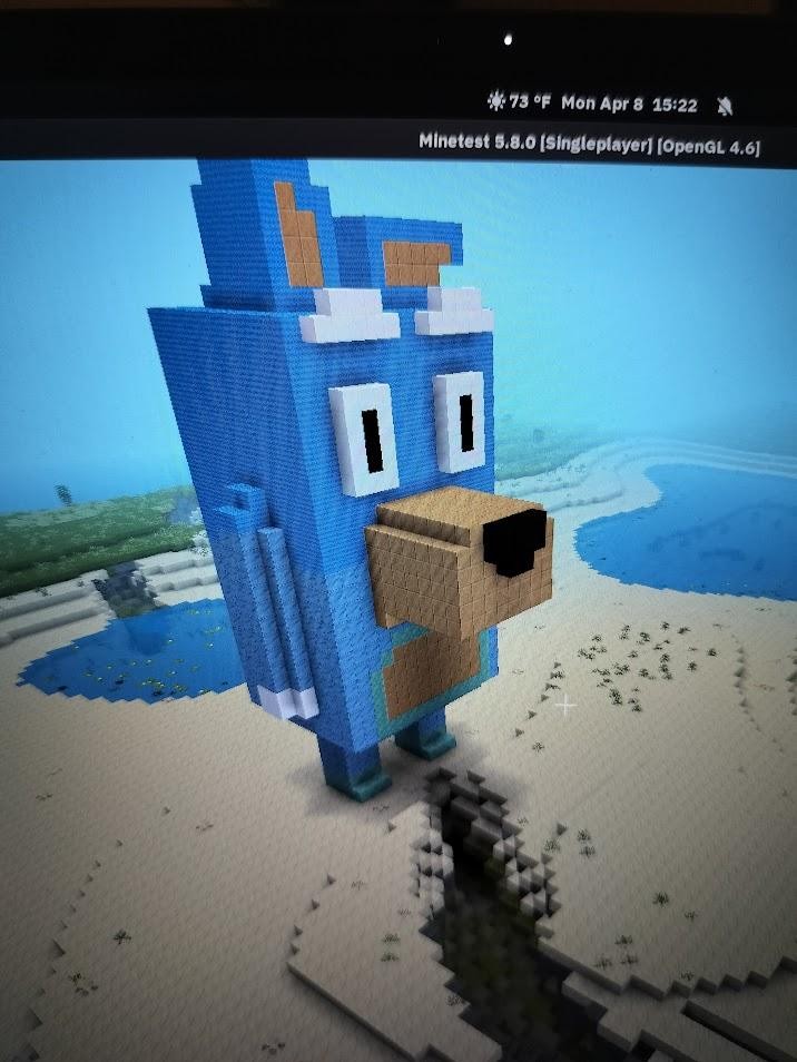 The old game Minetest, now renamed Luanti, running on GNU/Linux. On this level, there is a giant 3-D Bluey building standing on the sand. There is a lake in the background, and green grass. 