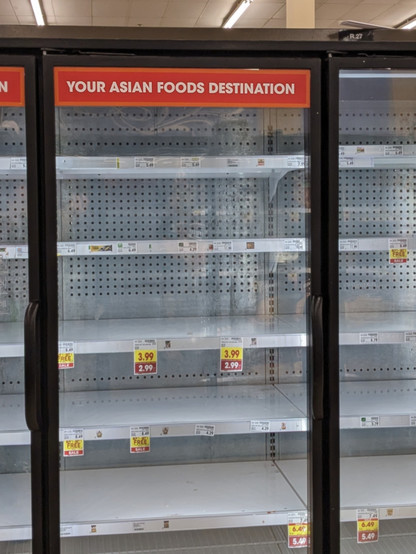 Shelves behind a glass door in a supermarket. There are a few price labels attached to the shelves, but the shelves are empty of any merchandise. A sign adhered to the top of the door reads in plain letters: "Your Asian Foods Destination". It's impossible to read the product names on the price labels. But two labels say $3.99 marked down to $2.99. Two others say $6.49 marked down to $5.49. And several labels say "Buy 1 get 1 free". The ceiling is barely visible in the photo past the shelves: a …