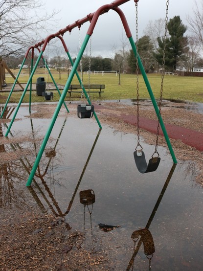 A children's swingset in a park that has high water. The swingset has a metal frame painted red and green, and three swings attached by chains. One is a plain flat black rubber swing, bowed down in the middle. The other two are black bucket seats with leg holes, allowing toddlers to swing without falling. There ground below the swingset is red mulch. However, the entire area of the swingset is covered in water. There reflections of each swing can be seen in the water, the chains going down to t…
