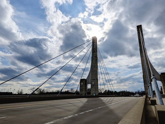 A multilane street going over a cable-stayed bridge. The bridge has a tower on each side of the road. Each tower has several support cables going from the top of tower down to the road. The sky is cloudy but the sun is trying to peek through , behind the left tower, making kind of a halo around the top of the tower, and casting a shadow from the tower onto the road.